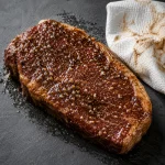 Daging sapi yang sudah dimarinasi dengan cairan bumbu gelap dan lada hitam di atas permukaan batu slate, dengan tisu dapur di sampingnya sebagai persiapan sebelum proses memasak steak