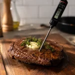 Steak yang sedang diistirahatkan di atas talenan kayu dengan termometer digital menunjukkan suhu 55 derajat Celcius, butter meleleh dan thyme segar di atasnya sebagai panduan cara masak steak medium rare yang benar